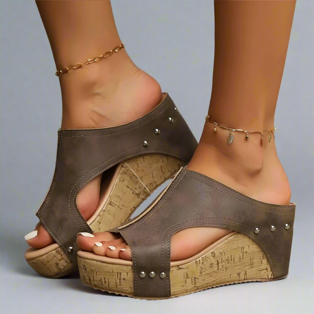 A pair of brown cork-wedge sandals with cutouts and stud accents, featuring a slip-on design and orthopedic footbed.
