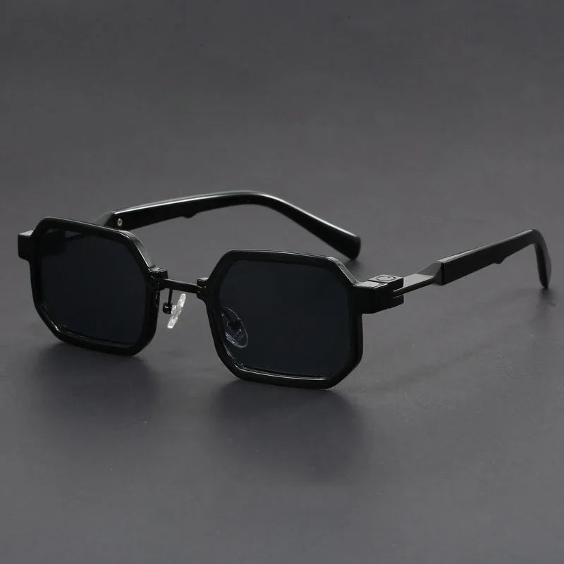 A pair of sunglasses with a unique octagonal frame, black with black lenses, displayed against a grey background.